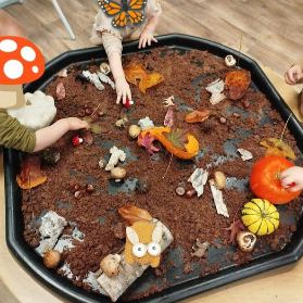 Un bac sensoriel d'automne rempli de terre, de feuilles et de figurines, exploré par les mains d'enfants.